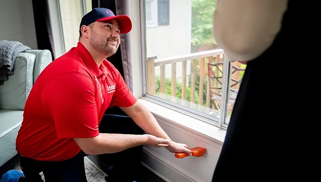 A Rainbow Restoration service professional bending down next to a wall inside a customer's home and gesturing to the reading on the moisture meter he's using below a large window.