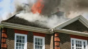 Smoke and flames rise from the roof of a brick home after a fire, showing why professional fire and smoke damage restoration may be needed.