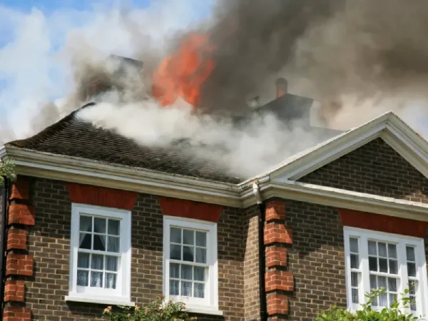 Smoke and flames rise from the roof of a brick home after a fire, showing why professional fire and smoke damage restoration may be needed.