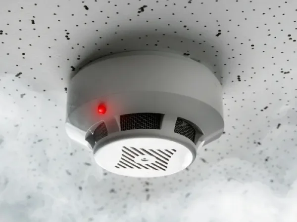A smoke detector with a red warning light is mounted on a speckled ceiling, representing the hidden smoke damage that can remain after a home fire.